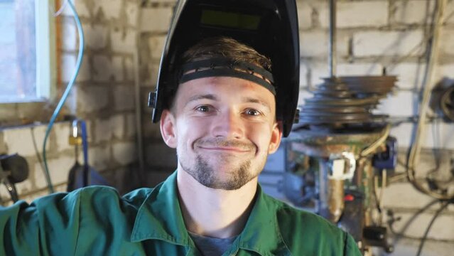 Happy young repairman opening protective mask and showing grimaces into camera. Portrait of mechanic in uniform making funny face and fooling around in garage. Welder with beard working in workshop