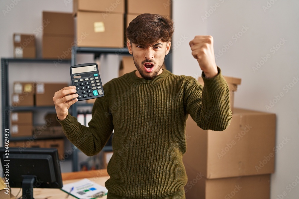 Poster arab man with beard working at small business ecommerce holding calculator annoyed and frustrated sh