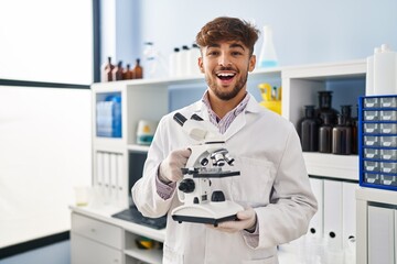 Arab man with beard working at scientist laboratory holding microscope smiling and laughing hard out loud because funny crazy joke.