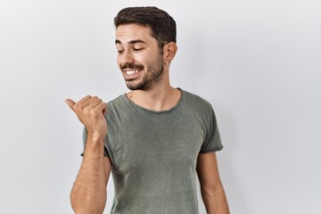 Fototapeta premium Young hispanic man with beard wearing casual t shirt over white background smiling with happy face looking and pointing to the side with thumb up.