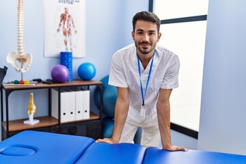 Young hispanic man wearing physiotherapist uniform leaning on massage table at rehab clinic - obrazy, fototapety, plakaty