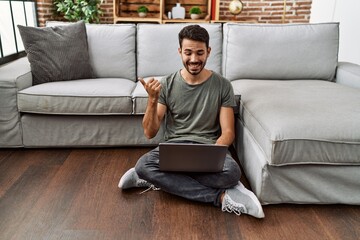 Young hispanic man using laptop sitting on the at home pointing thumb up to the side smiling happy with open mouth