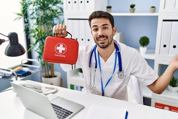 Young hispanic doctor man holding first aid kit celebrating achievement with happy smile and winner expression with raised hand