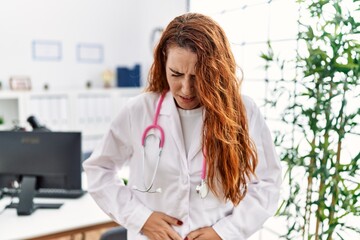 Young redhead woman wearing doctor uniform and stethoscope at the clinic with hand on stomach because indigestion, painful illness feeling unwell. ache concept.