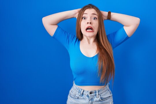 Redhead woman standing over blue background crazy and scared with hands on head, afraid and surprised of shock with open mouth
