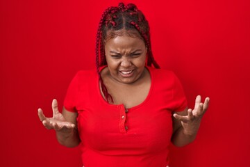 African american woman with braided hair standing over red background crazy and mad shouting and yelling with aggressive expression and arms raised. frustration concept. - obrazy, fototapety, plakaty