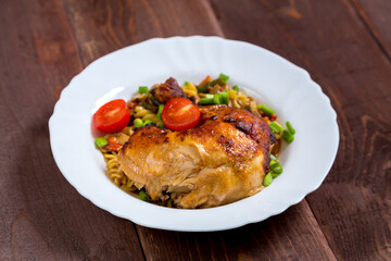 Pasta with vegetables and baked chicken leg sprinkled with green onions in a plate on a wooden table.