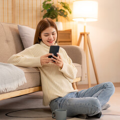 Young asian woman chatting with customer on smartphone and drinking coffee while wearing sweater and