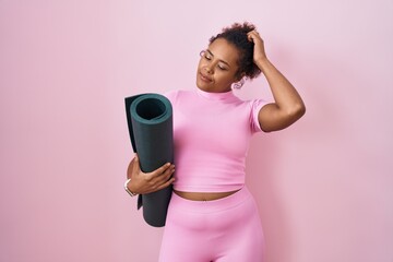 Young hispanic woman with curly hair holding yoga mat over pink background confuse and wondering...