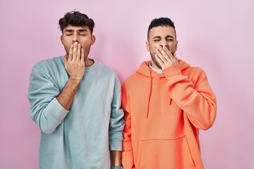 Young hispanic gay couple standing over pink background bored yawning tired covering mouth with hand. restless and sleepiness.