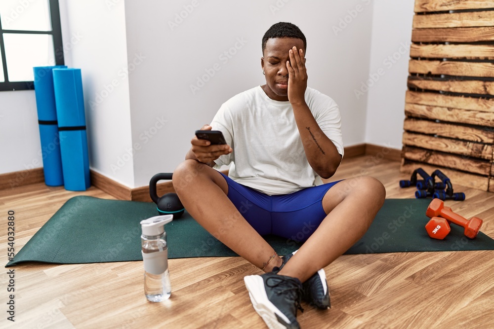 Wall mural young african man sitting on training mat at the gym using smartphone yawning tired covering half fa