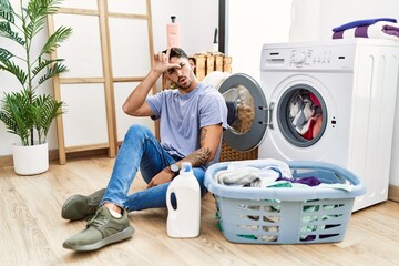 Young hispanic man putting dirty laundry into washing machine making fun of people with fingers on forehead doing loser gesture mocking and insulting.