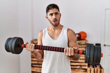 Young hispanic man training with weighs in shock face, looking skeptical and sarcastic, surprised...