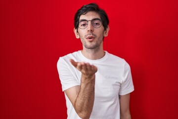 Young hispanic man standing over red background looking at the camera blowing a kiss with hand on air being lovely and sexy. love expression.