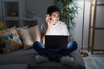 Young hispanic man using laptop at home at night smiling with hand over ear listening an hearing to rumor or gossip. deafness concept.
