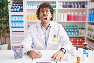 Hispanic young man working at pharmacy drugstore angry and mad screaming frustrated and furious, shouting with anger. rage and aggressive concept.