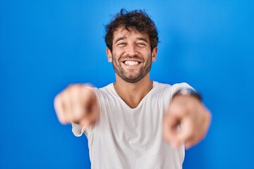 Hispanic young man standing over blue background pointing to you and the camera with fingers, smiling positive and cheerful