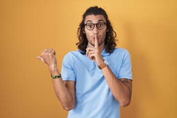 Young hispanic man standing over yellow background asking to be quiet with finger on lips pointing with hand to the side. silence and secret concept.