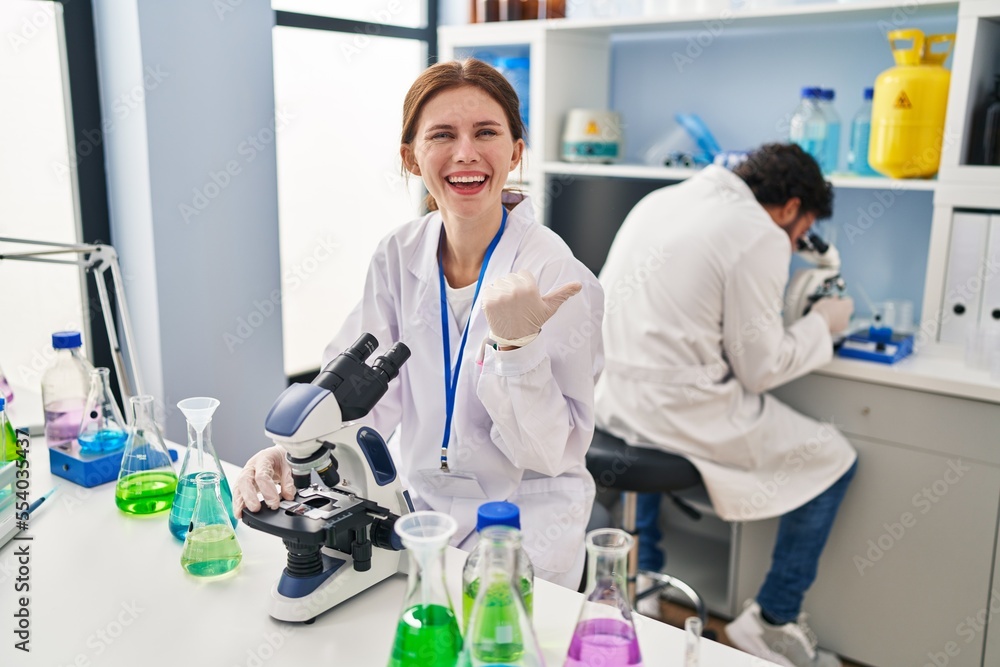 Poster young two people working at scientist laboratory pointing thumb up to the side smiling happy with op