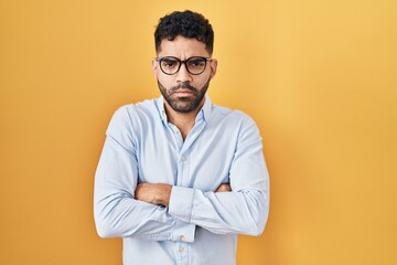 Hispanic man with beard standing over yellow background skeptic and nervous, disapproving expression on face with crossed arms. negative person.