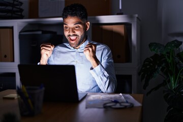 Hispanic man with beard working at the office with laptop at night screaming proud, celebrating victory and success very excited with raised arms