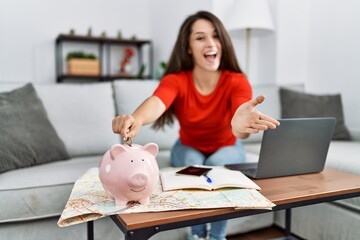 Obraz na płótnie Canvas Young brunette woman putting euro coin in piggy bank saving for travel celebrating achievement with happy smile and winner expression with raised hand