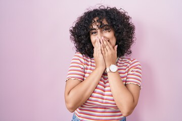 Young middle east woman standing over pink background laughing and embarrassed giggle covering...