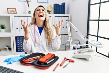 Young beautiful doctor woman with reflex hammer and medical instruments crazy and mad shouting and...