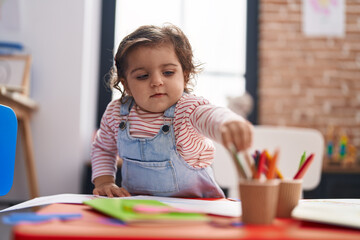 Adorable hispanic girl student sitting on table drawing on paper at kindergarten