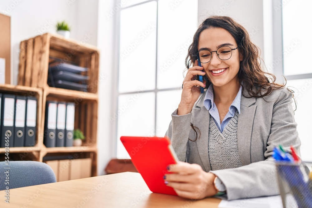 Wall mural Young hispanic woman business worker using touchpad and talking on the smartphone at office