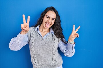 Young brunette woman standing over blue background smiling with tongue out showing fingers of both hands doing victory sign. number two.