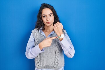 Young brunette woman standing over blue background in hurry pointing to watch time, impatience, looking at the camera with relaxed expression