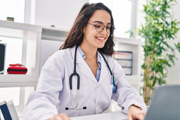 Young hispanic woman wearing doctor uniform using laptop working at clinic