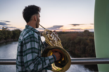 Young man playing his golden horn on a bridge at sunset.
