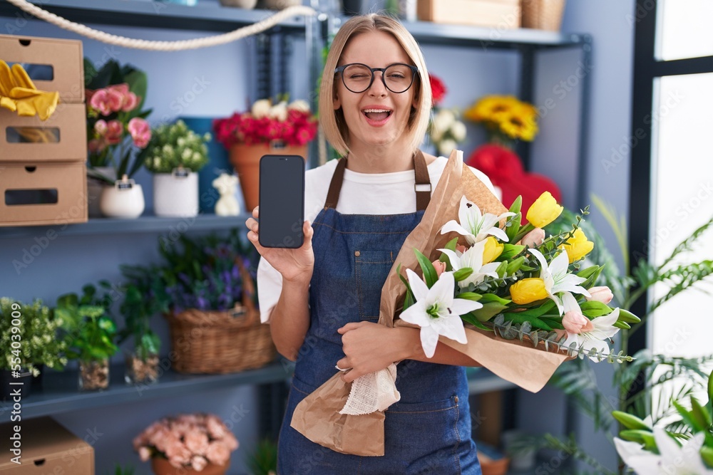 Wall mural young caucasian woman working at florist shop showing smartphone screen winking looking at the camer