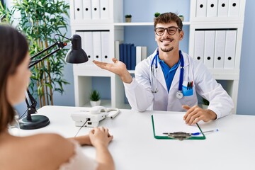 Young doctor with client at medical clinic smiling cheerful presenting and pointing with palm of hand looking at the camera.