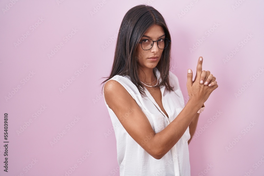 Sticker Brunette young woman standing over pink background wearing glasses holding symbolic gun with hand gesture, playing killing shooting weapons, angry face