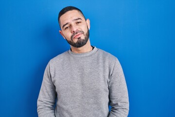 Hispanic man standing over blue background with hands together and crossed fingers smiling relaxed and cheerful. success and optimistic