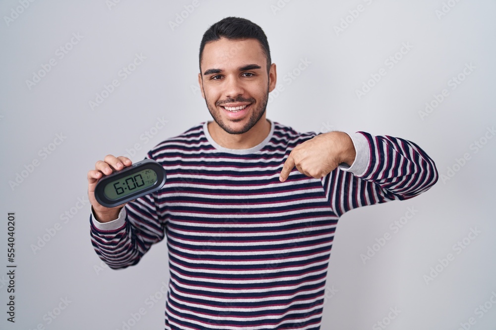 Poster Young hispanic man wearing pyjama holding alarm clock pointing finger to one self smiling happy and proud