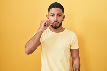 Young hispanic man standing over yellow background mouth and lips shut as zip with fingers. secret and silent, taboo talking