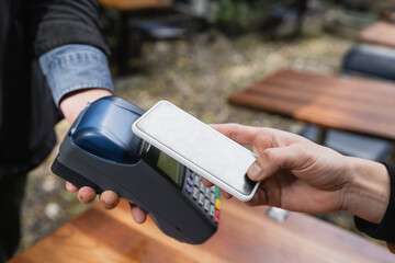 Cropped view of man paying with cellphone near waitress with credit card reader in outdoor cafe.