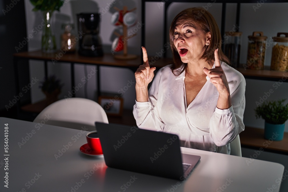 Sticker middle age hispanic woman using laptop at home at night amazed and surprised looking up and pointing