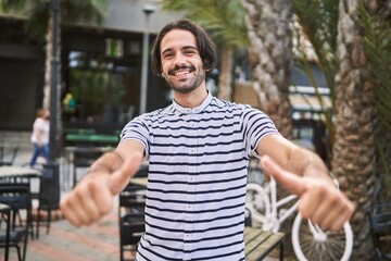 Young hispanic man with beard outdoors at the city approving doing positive gesture with hand, thumbs up smiling and happy for success. winner gesture.