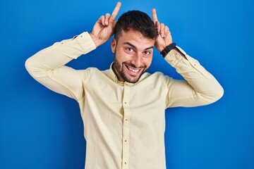 Handsome hispanic man standing over blue background posing funny and crazy with fingers on head as bunny ears, smiling cheerful