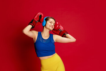 Portrait of young beautiful girl in sportive clothes posing in headphones and boxing gloves over red background