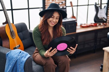 Young hispanic woman musician holding vinyl disc sitting on sofa at music studio