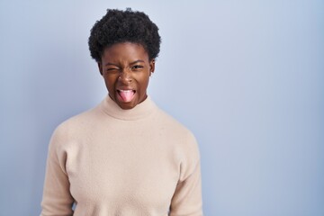 African american woman standing over blue background sticking tongue out happy with funny...