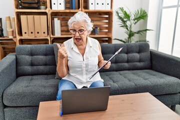 Senior woman with grey hair working at consultation office doing online therapy pointing thumb up to the side smiling happy with open mouth