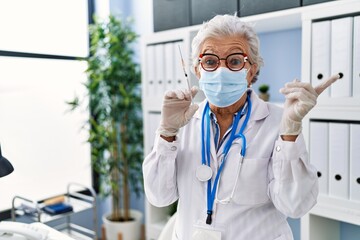 Fototapeta premium Senior woman with grey hair wearing doctor uniform and medical mask holding syringe smiling happy pointing with hand and finger to the side