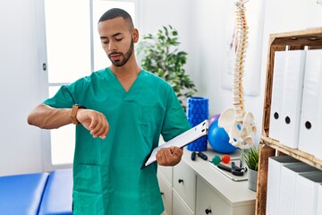 African american physiotherapist man working at pain recovery clinic checking the time on wrist...